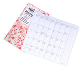 A mini monthly planner shown open to July 2028, with a floral-patterned cover underneath and a white interior page displaying the calendar layout. The cover reads &