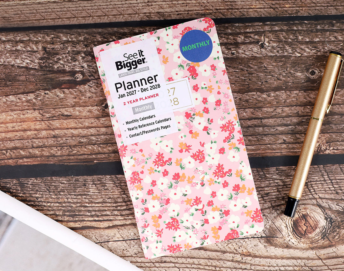 A floral monthly pocket planner by See It Bigger lies on a rustic wooden tabletop next to a gold pen. The planner cover features a light pink background with a red, orange, and green floral pattern, and includes a blue circular &