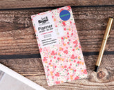 A floral monthly pocket planner by See It Bigger lies on a rustic wooden tabletop next to a gold pen. The planner cover features a light pink background with a red, orange, and green floral pattern, and includes a blue circular &